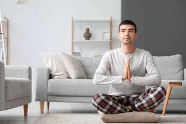 Young man in pajamas meditating on pillow at home