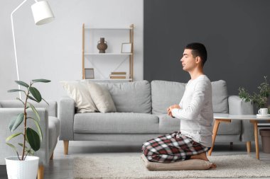 Young man in pajamas meditating on pillow at home