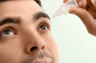 Young man using eye drops on green background, closeup