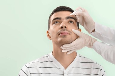 Ophthalmologist putting drops in young man's eye on green background