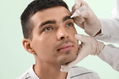 Ophthalmologist putting drops in young man's eye on green background, closeup