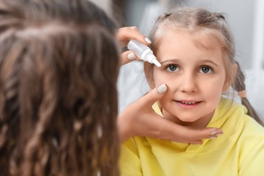 Mother putting drops in her little daughter's eye at home, closeup