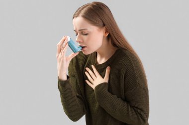 Sick young woman with inhaler on grey background