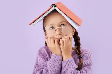 Little girl with book biting nails on lilac background, closeup