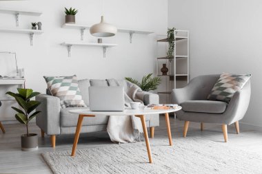Cozy interior of living room with grey sofa, armchair and modern laptop on table