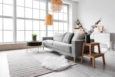 Cozy interior of living room with grey sofa, pouf and fur carpet near window