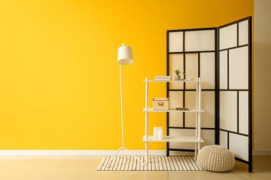 Interior of room with folding screen, shelving unit and pouf near yellow wall