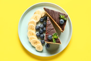 Plate with pieces of birds milk cake, blueberries, banana slices and mint leaves on yellow background