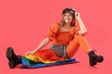 Beautiful young woman in hat with LGBT flag sitting on red background