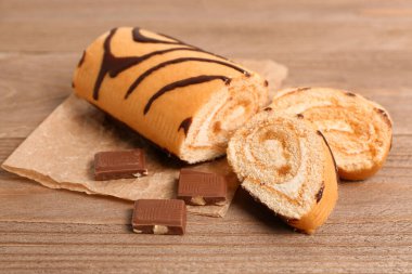 Baking paper with pieces of delicious sponge cake roll and chocolate on wooden table