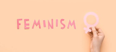 Female hand with paper symbol of woman and word FEMINISM on beige background