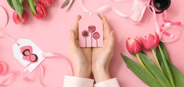 Female hands with paper tag for decorating gift boxes, tulip flowers and ribbon on pink background. International Women's Day celebration