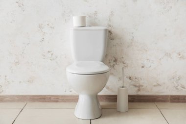 Ceramic toilet bowl with paper roll and brush near light wall