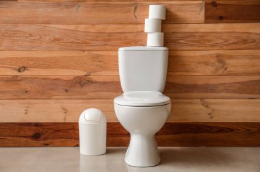 Ceramic toilet bowl with paper rolls and bin near wooden wall