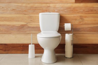 Ceramic toilet bowl, holder with paper rolls and brush near wooden wall
