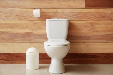 Ceramic toilet bowl, paper roll and bin near wooden wall