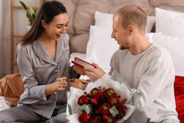 Young man greeting his wife with jewelry in bedroom on Valentine's Day