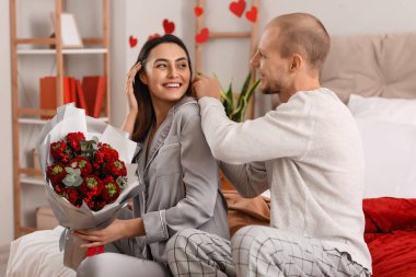 Young man putting necklace around his wife's neck in bedroom on Valentine's Day