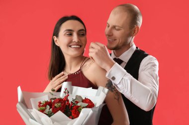 Young man putting necklace around his girlfriend's neck on red background. Valentine's Day celebration