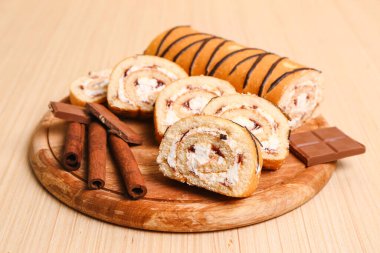 Cutting board with tasty pieces of sponge cake roll, cinnamon and chocolate on wooden background
