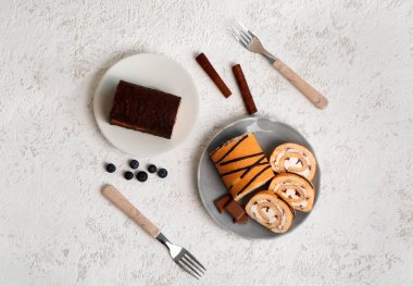 Plates with tasty sponge cake roll, cinnamon, coffee beans, chocolate and forks on light grunge background
