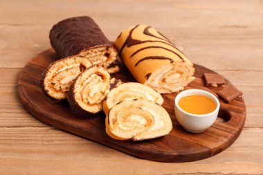 Board with pieces of delicious sponge cake roll, bowl of jam and chocolate on wooden table