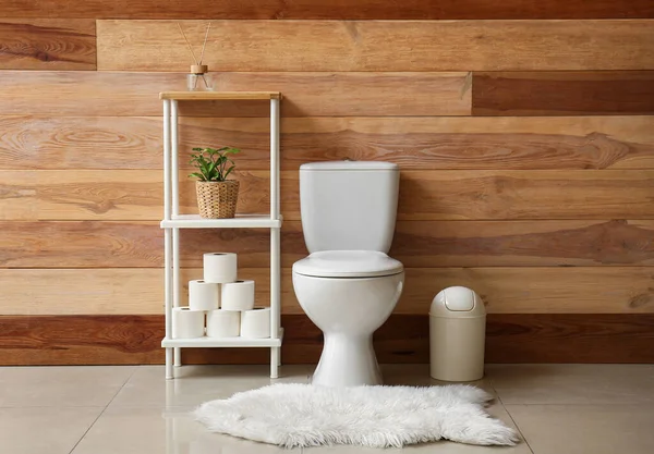 Ceramic toilet bowl, bin and shelving unit with paper rolls near wooden wall