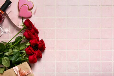 Bottle of wine, rose flowers, gift and heart-shaped cookies on pink tile table. Valentine's Day celebration