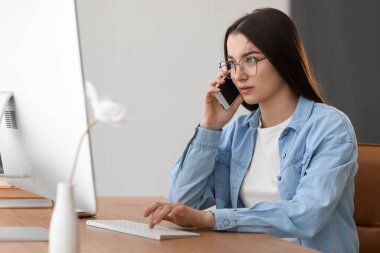 Young woman talking by mobile phone and using computer at table in office
