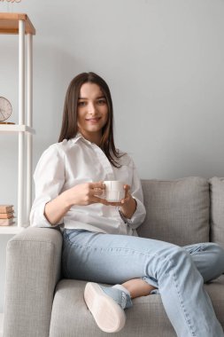 Evdeki kanepede oturan bir fincan sıcak içecekle genç bir kadın.