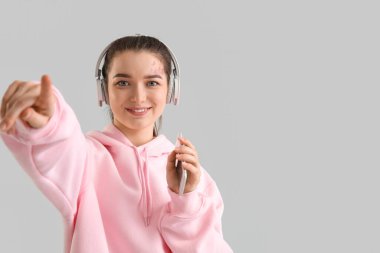 Young woman in headphones with mobile phone pointing at viewer on light background
