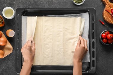 Woman with dough, parchment paper, baking tray and ingredients on dark background