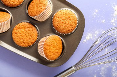 Baking tray with muffins, whisk and scattered flour on lilac background