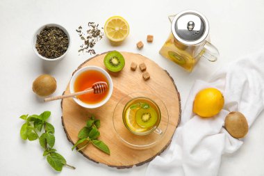 Board with glass cup of fruit tea, kiwi, lemon, mint, honey and teapot on white background