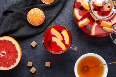 Glass cup of fruit tea with grapefruit, mandarin, teapot and napkin on dark background