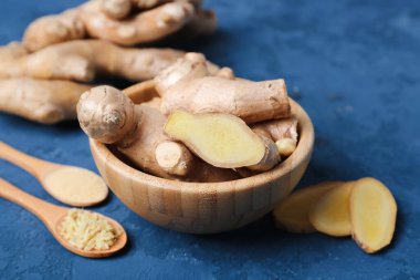 Wooden bowl with fresh ginger roots, slices and spoons of powder on blue grunge background, closeup