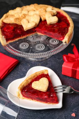 Heart-shaped plate with piece of sweet strawberry pie, cookie hearts and gift for Valentine's day on dark table
