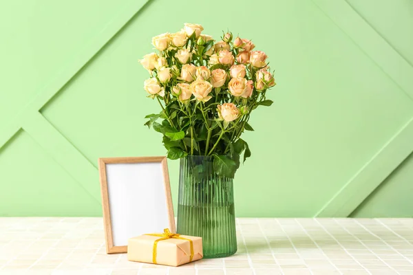 Vase with beautiful rose flowers, empty picture frame and gift box on table near color wall