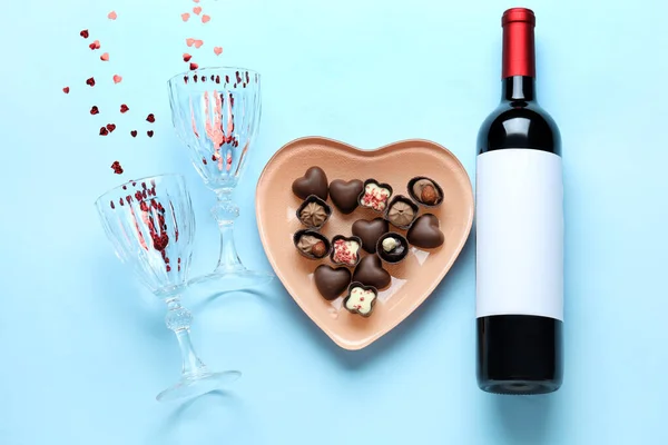 Bottle of wine, plate with chocolate candies and glasses on blue background. Valentine\'s Day celebration