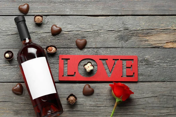 Bottle of wine, chocolate candies, word love and rose flower on wooden background. Valentine\'s Day celebration