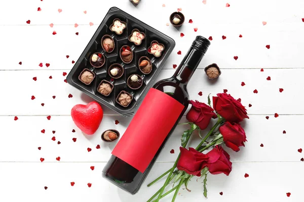 Bottle of wine, box with chocolate candies and rose flowers on white wooden table. Valentine\'s Day celebration