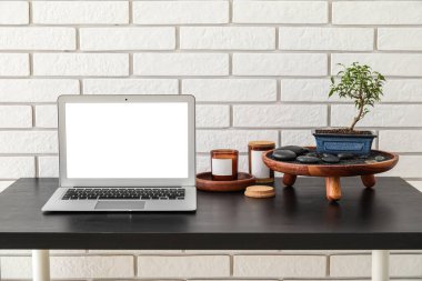Stand with Bonsai tree, spa stones, candles and laptop on table near white brick wall
