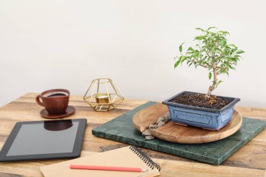 Table with Bonsai tree, tablet computer, cup of beverage and notebook near light wall