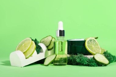 Cosmetic dropper bottle with jar of cream, green moss, cucumber slices and lime on color background