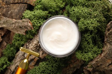 Jar of cream with facial massage tool and green moss on tree bark, closeup