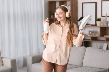 Young woman in headphones with rag and detergent dancing at home