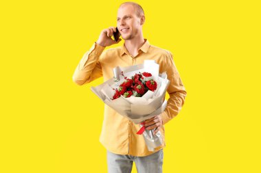 Young man with bouquet of flowers talking by mobile phone on yellow background. Valentine's Day celebration