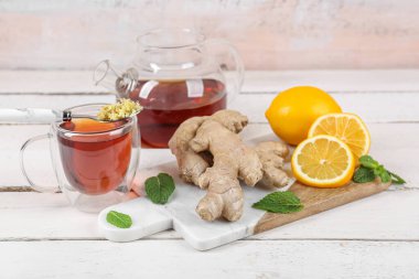 Board with ginger roots, lemon, glass cup of tea and teapot on white wooden table