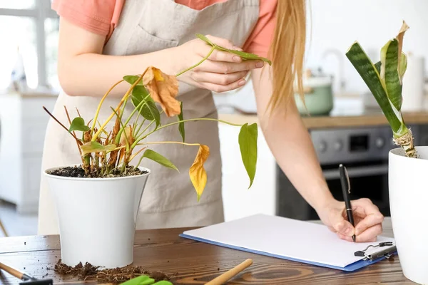 Woman with wilted houseplant writing in clipboard at home, closeup