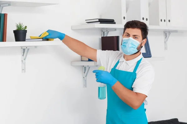Male janitor with medical mask cleaning bookshelf in office